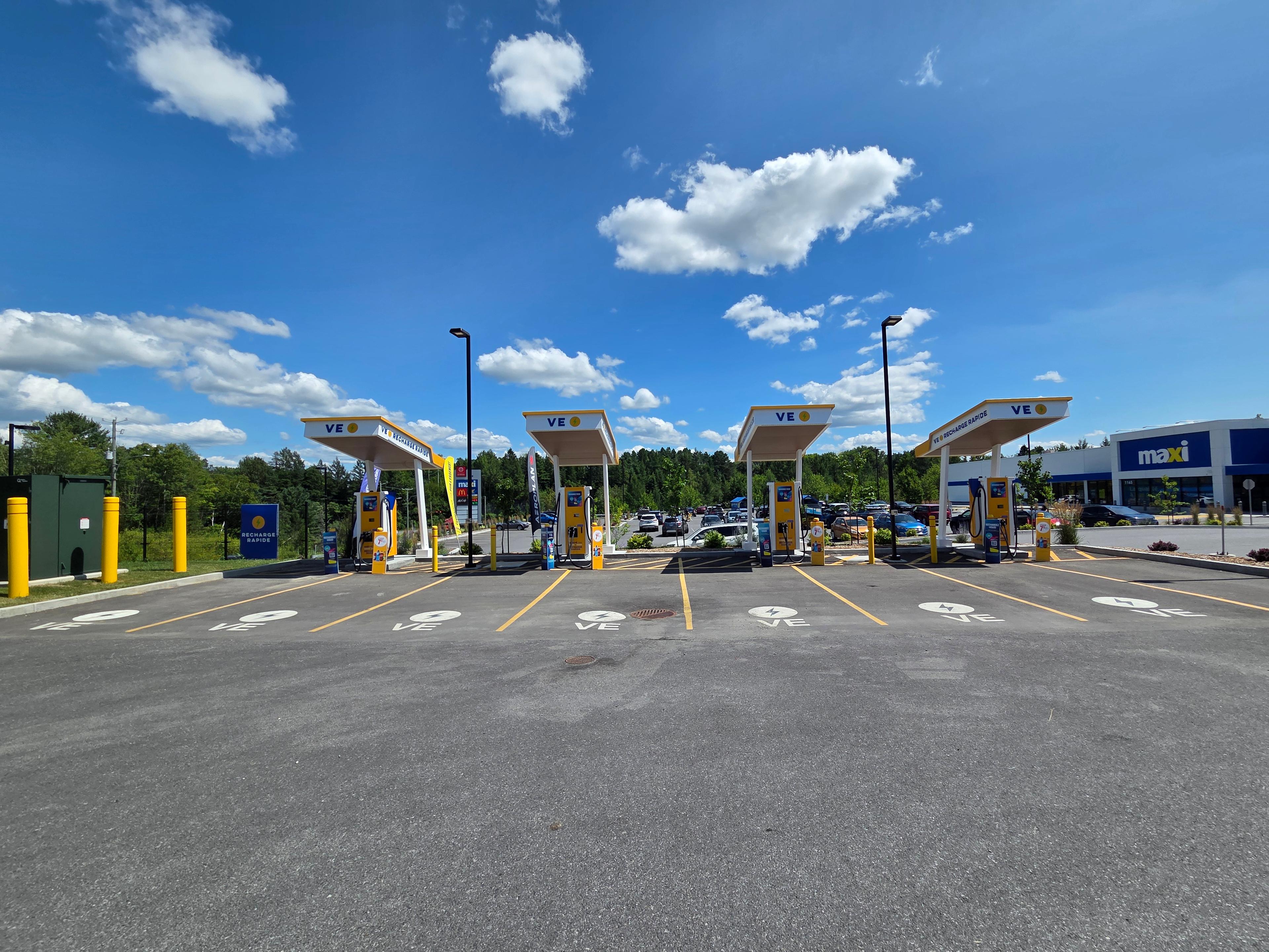 The photo shows a VE Recharge Rapide station with four yellow and blue fast chargers under white canopies. The site is clearly marked for EVs, with painted VE symbols on the parking spots. A Maxi grocery store is visible in the background.