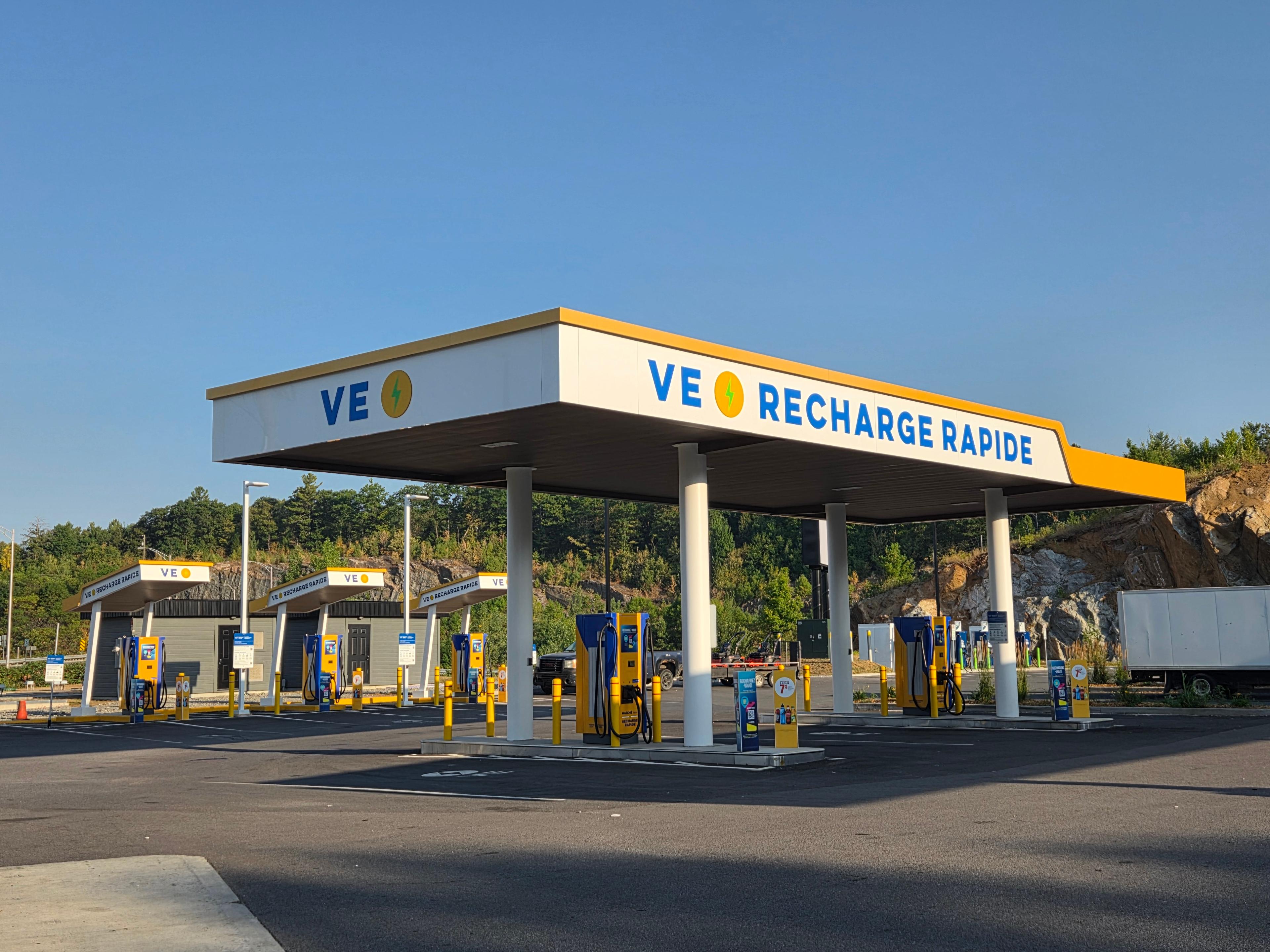 The photo shows a VE Recharge Rapide station with multiple yellow and blue charging units under a large white canopy. The signage prominently displays “VE Recharge Rapide” in blue letters with a yellow lightning bolt icon. The site is clean and spacious, with several chargers lined up and clear parking markings.